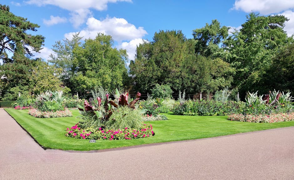 A meticulously maintained garden border featuring vibrant, well-pruned plants and flower beds arranged along a curved pathway. The border includes large, sculptural plants with broad, dark green and r