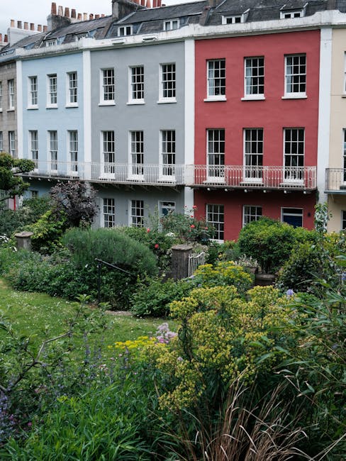 A well-maintained front garden scene in Teddington during daytime, featuring a neatly mown green lawn bordered by garden beds filled with a variety of flowering plants and shrubs. There is a paved pat